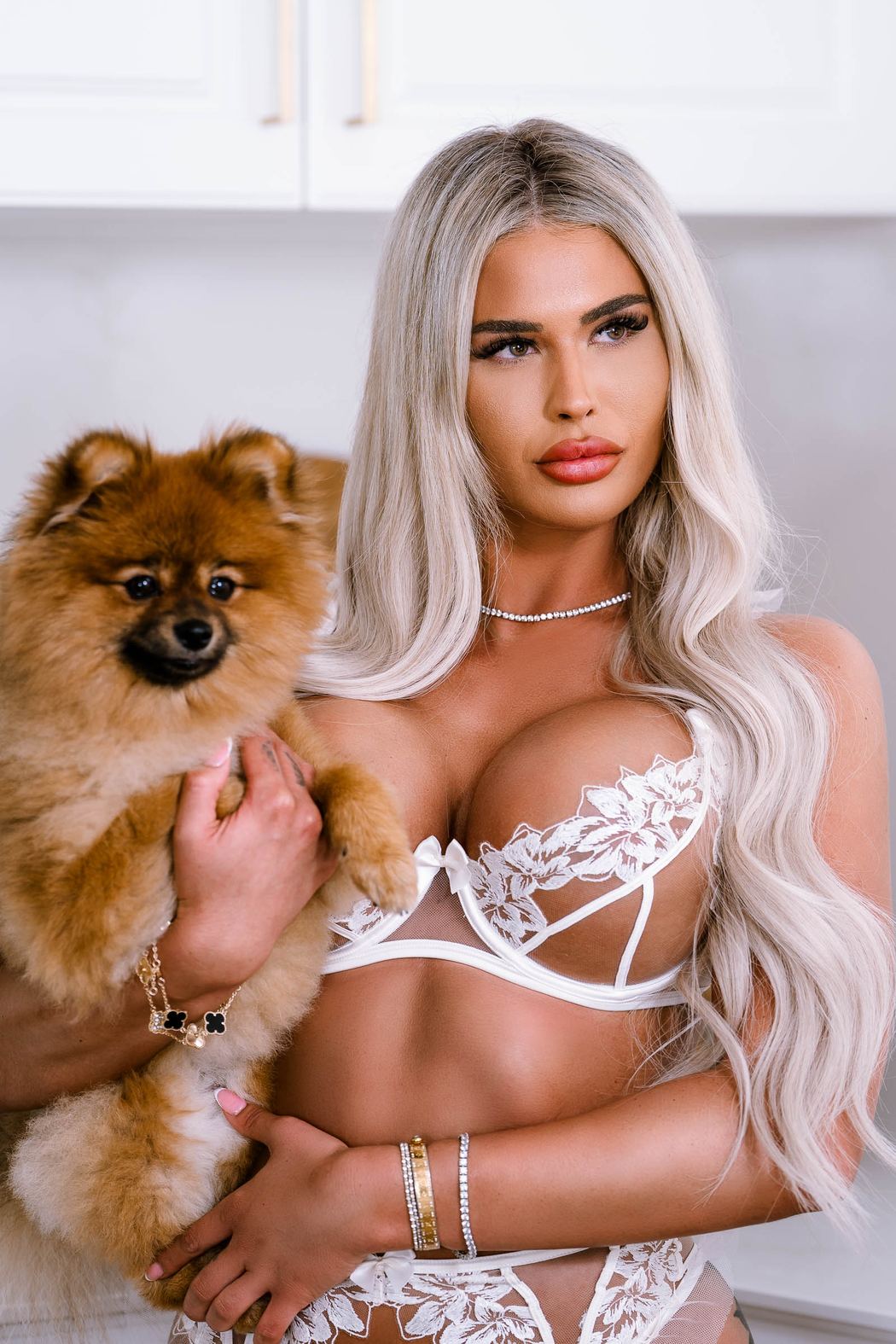 Woman in white lace lingerie holding a small dog in a bright kitchen